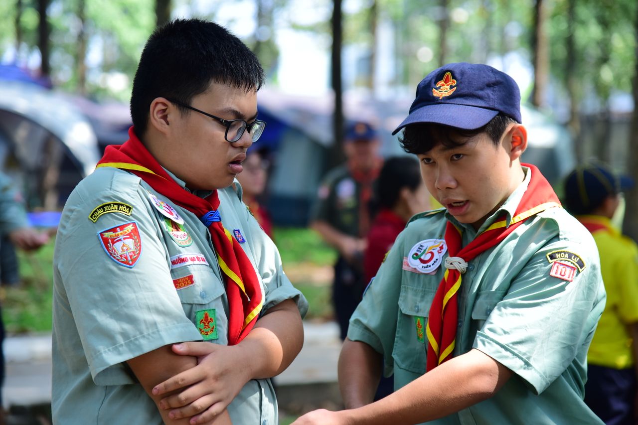Two boy scouts in uniform interact during an outdoor camping session in a forested area.