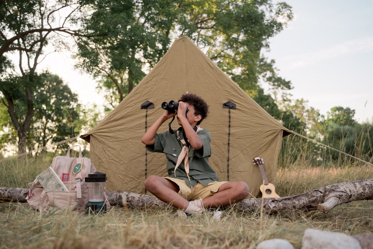 A young scout sits outside a tent, using binoculars to explore nature, surrounded by camping gear.