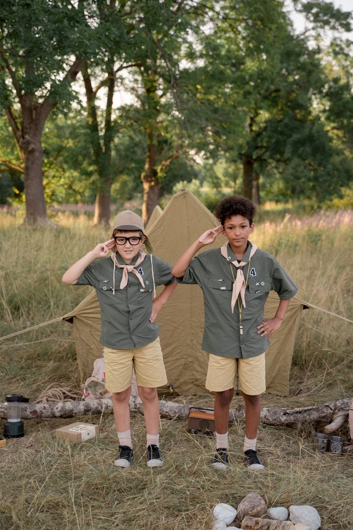 Two boys in scout uniforms saluting in front of a tent during a sunny day camp.